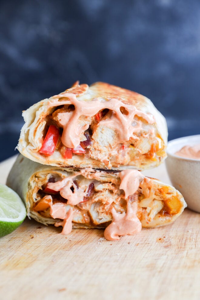 A close-up of a grilled chicken fajitas wrap cut in half, revealing chicken, peppers, and sauce inside. Creamy pink sauce drips from the filling. A lime wedge and a small bowl of sauce are beside it on a wooden surface.