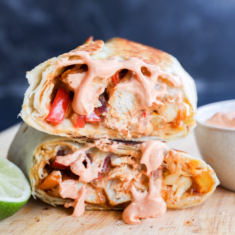 A close-up of a grilled chicken fajitas wrap cut in half, revealing chicken, peppers, and sauce inside. Creamy pink sauce drips from the filling. A lime wedge and a small bowl of sauce are beside it on a wooden surface.