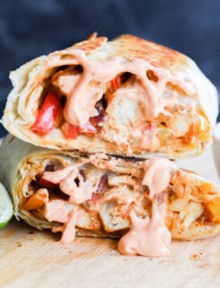 Two halves of a grilled sandwich, stuffed with vegetables and chicken, topped with creamy pink sauce, sit on a wooden board next to a halved lime and a small bowl of extra sauce.
