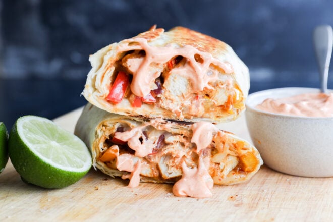 Two halves of a grilled sandwich, stuffed with vegetables and chicken, topped with creamy pink sauce, sit on a wooden board next to a halved lime and a small bowl of extra sauce.