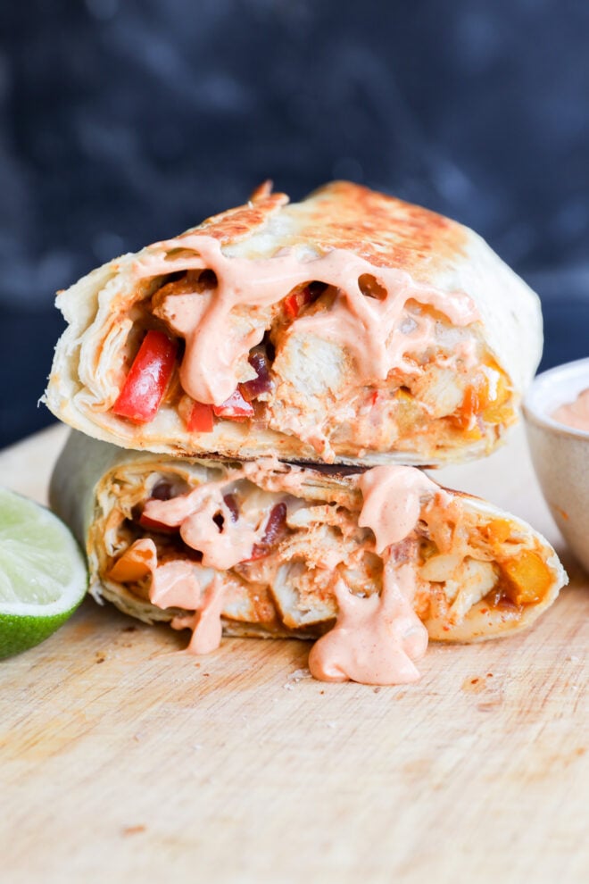 Two halves of a chicken fajitas wrap filled with grilled chicken, red peppers, onions, and melted cheese, topped with creamy sauce, displayed on a wooden board with a lime wedge and a small bowl of sauce nearby.