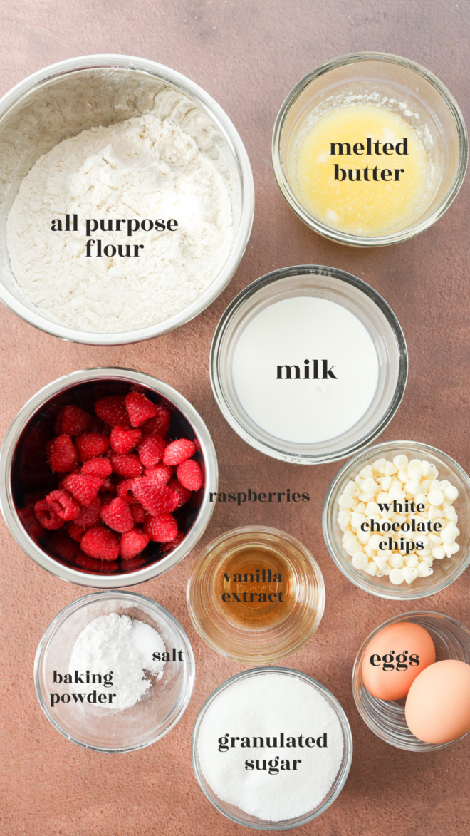 Top-down view of labeled baking ingredients for Raspberry White Chocolate Muffins: all-purpose flour, melted butter, milk, fresh raspberries, white chocolate chips, vanilla extract, eggs, sugar, salt, and baking powder.