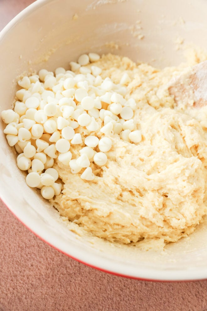 A mixing bowl filled with batter and a pile of white chocolate chips on top, partially mixed in with a wooden spoon.