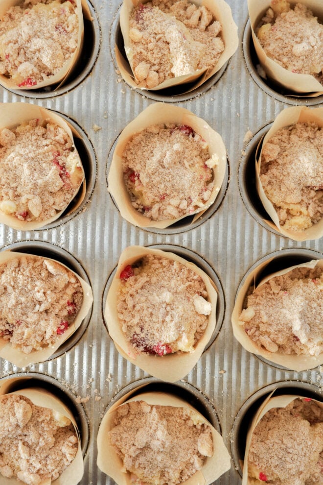 Twelve unbaked Raspberry White Chocolate Muffins in parchment paper liners sit in a metal muffin tray, topped with a crumbly streusel mixture, ready to be baked.