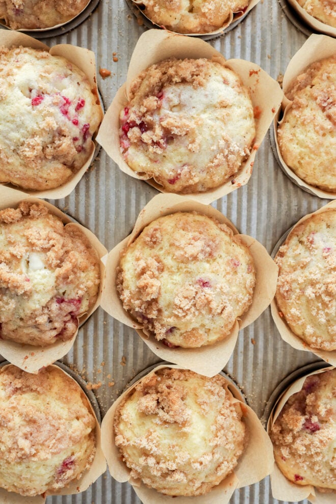 Overhead view of several freshly baked Raspberry White Chocolate Muffins in brown paper wrappers, arranged closely on a baking tray. The muffins have a crumbly topping and bits of red fruit are visible in some.