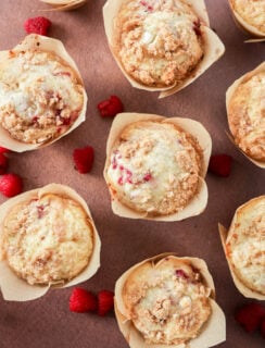 Overhead view of several Raspberry White Chocolate Muffins in parchment paper wrappers, arranged on a pink surface with scattered fresh raspberries. The muffins have crumbly tops and visible bits of raspberry.