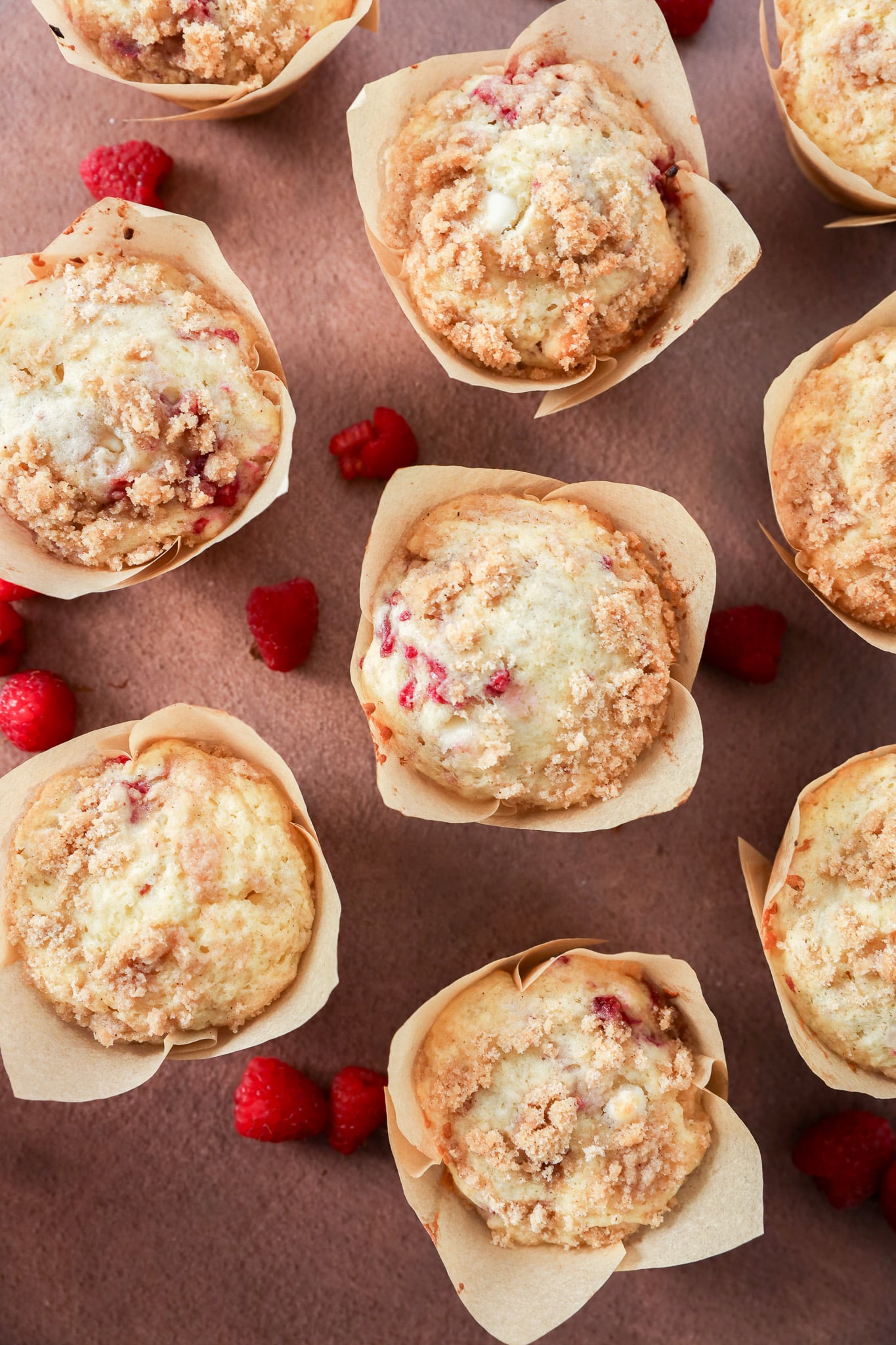 Overhead view of several Raspberry White Chocolate Muffins in parchment paper wrappers, arranged on a pink surface with scattered fresh raspberries. The muffins have crumbly tops and visible bits of raspberry.