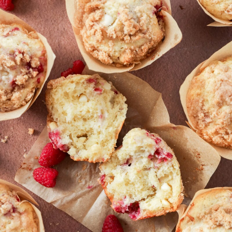 Overhead view of Raspberry White Chocolate Muffins, some whole and one split in half, revealing a moist interior studded with raspberries and white chocolate chips. Fresh raspberries are scattered on the brown surface around the muffins.