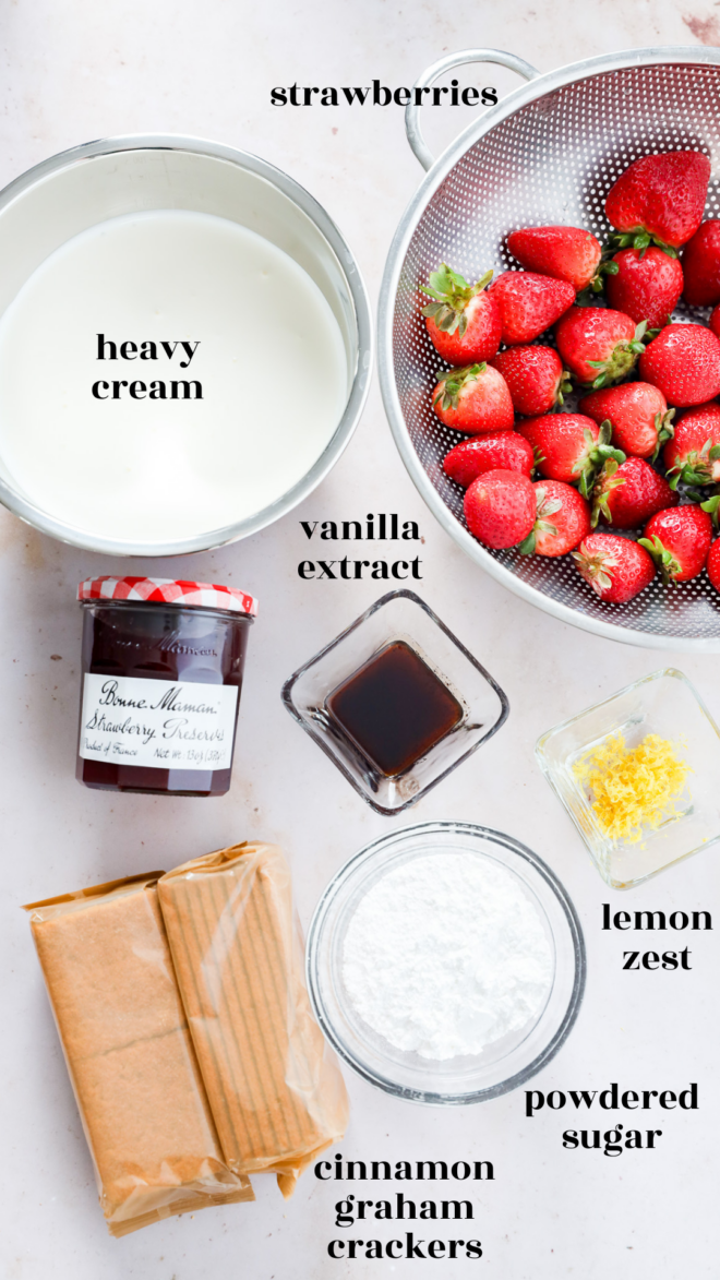 Overhead view of ingredients on a light surface: heavy cream in a bowl, fresh strawberries in a colander, vanilla extract, lemon zest, powdered sugar, cinnamon graham crackers, and a jar of strawberry preserves for strawberry icebox cake.