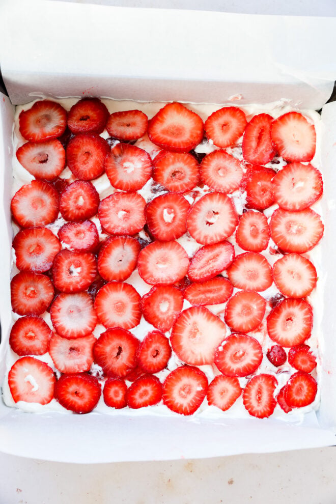 A square pan filled with a strawberry icebox cake, topped with neatly arranged, fresh strawberry slices on a layer of white cream.