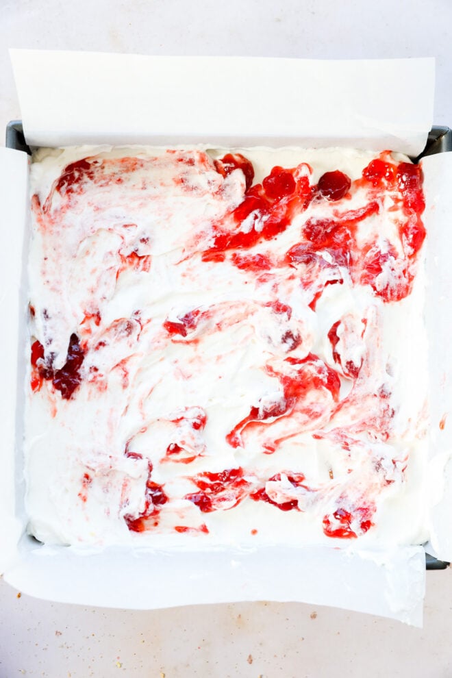 A square baking pan lined with parchment paper, filled with a creamy mixture and swirled red strawberry sauce on top,.