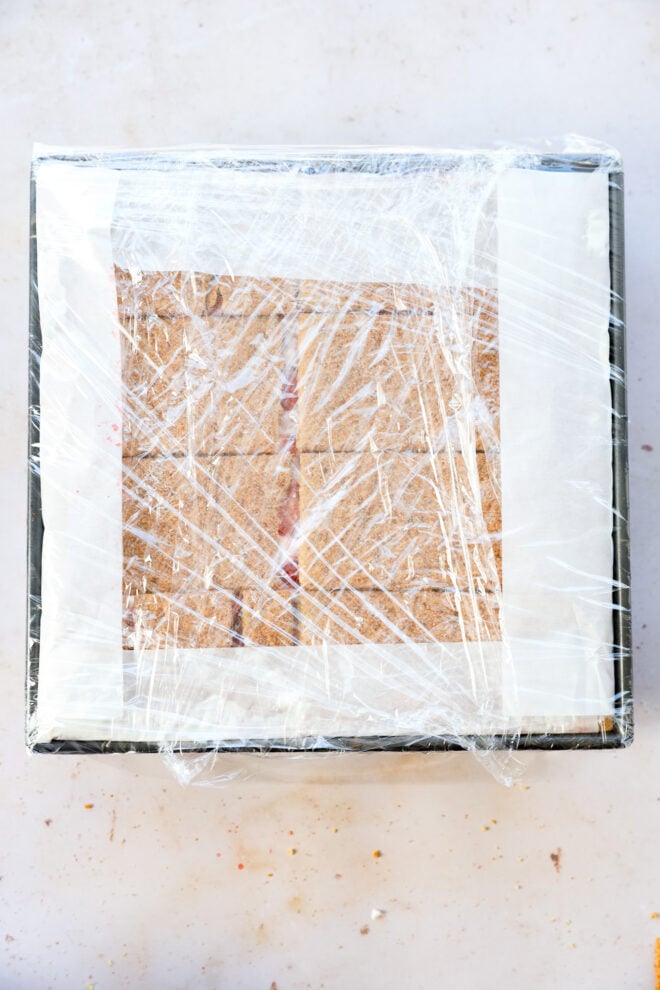 A square baking pan lined with parchment paper holds a pressed mixture for a strawberry icebox cake, covered tightly with plastic wrap. The pan rests on a light-colored surface.