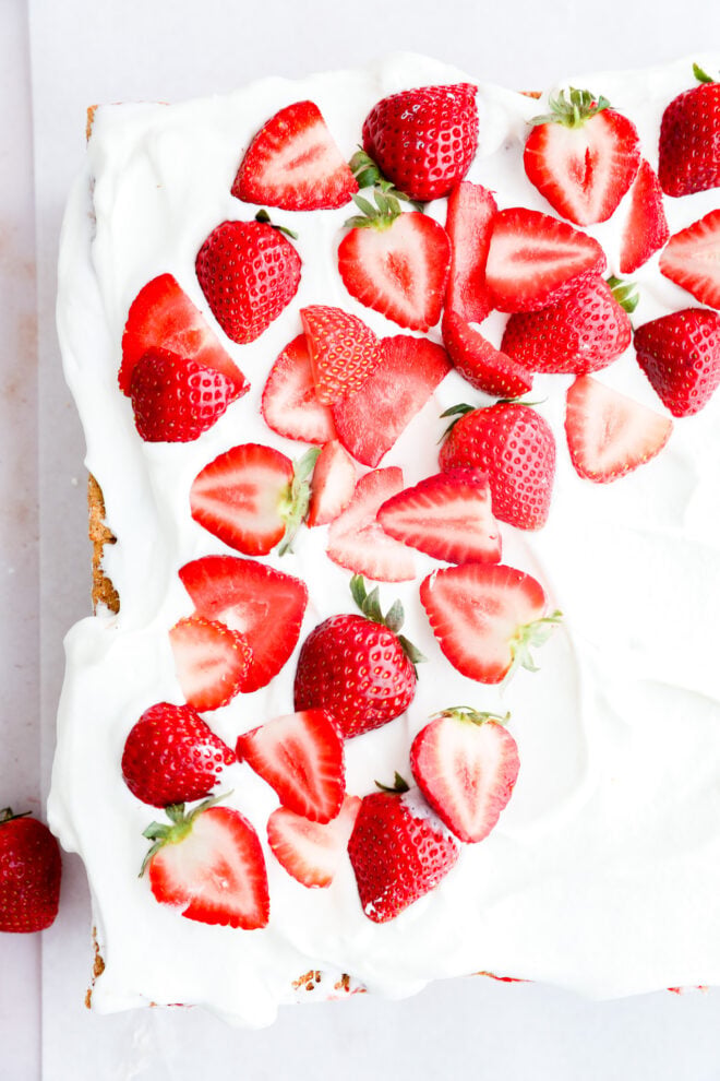 A rectangular strawberry icebox cake topped with a thick layer of whipped cream and fresh, halved strawberries, seen from above on a white surface.