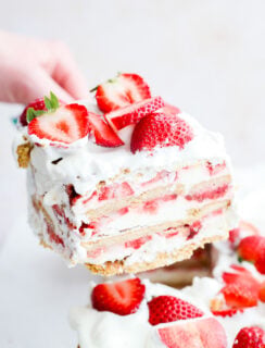 A hand lifts a slice of layered strawberry shortcake with whipped cream and fresh strawberry slices, revealing creamy and fruity layers reminiscent of a classic strawberry icebox cake.