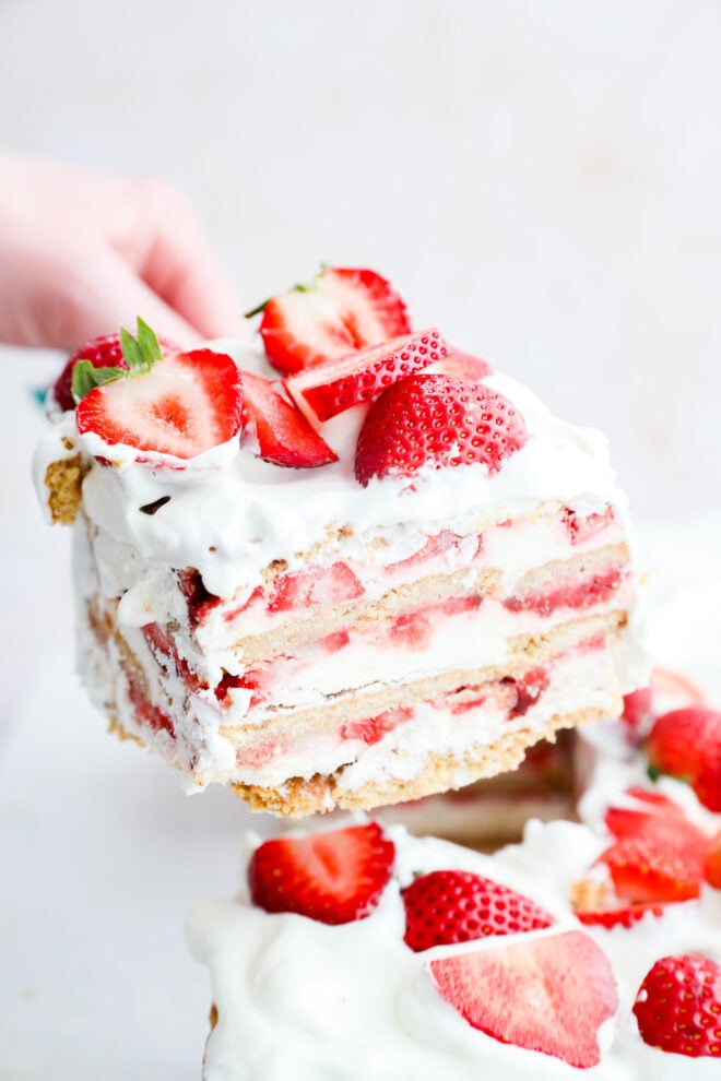 A hand lifts a slice of layered strawberry shortcake with whipped cream and fresh strawberry slices, revealing creamy and fruity layers reminiscent of a classic strawberry icebox cake.
