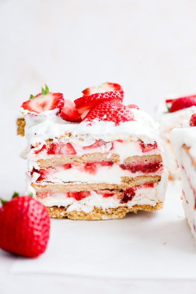 A close-up of a no-bake summer dessert shows its layers of whipped cream, graham crackers, and fresh fruit slices on top. A whole strawberry sits next to the cake slice on a white surface.