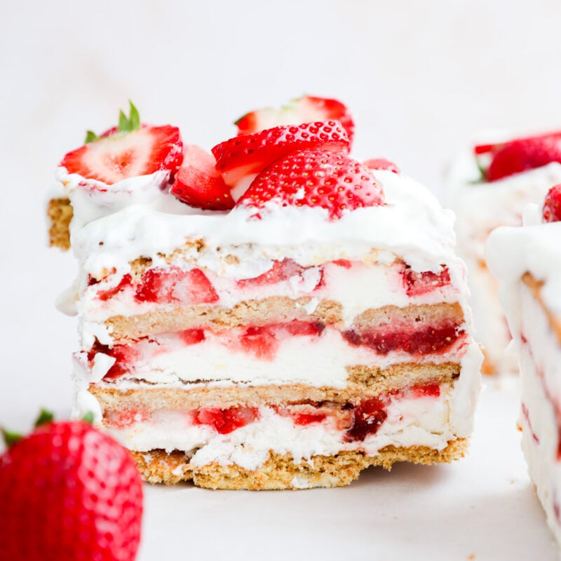 A close-up of a no-bake summer dessert shows its layers of whipped cream, graham crackers, and fresh fruit slices on top. A whole strawberry sits next to the cake slice on a white surface.