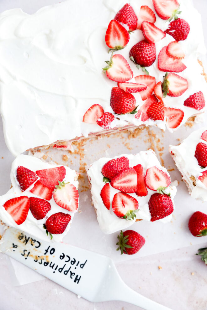A strawberry icebox cake topped with whipped cream and fresh strawberry halves, with two slices cut out. A cake server with the words Happiness is a piece of cake rests beside it on a white surface.