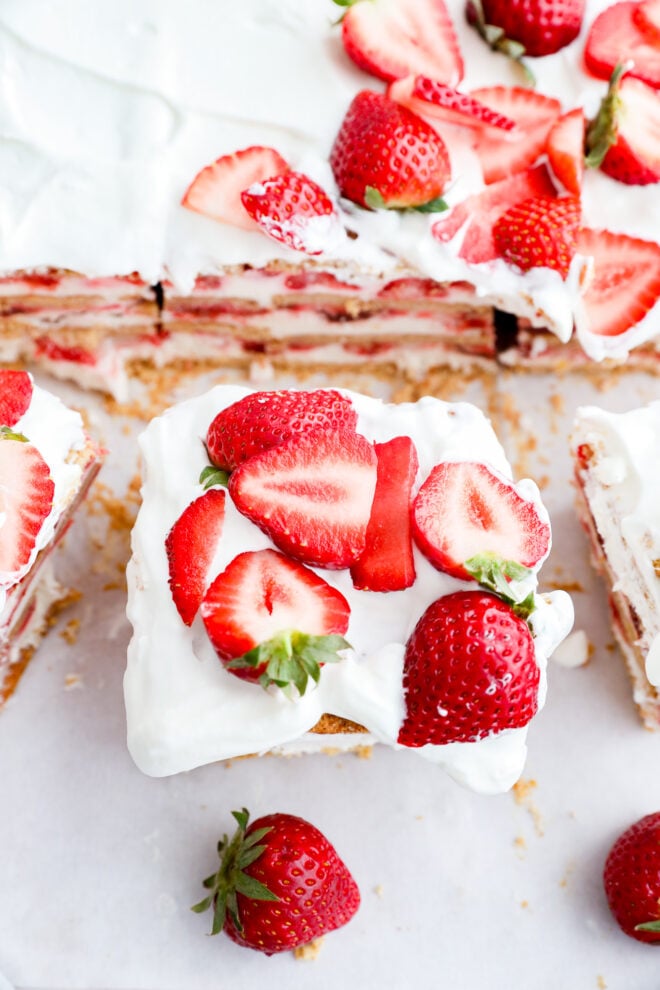 A slice of a no-bake summer dessert topped with whipped cream and fresh, sliced strawberries sits on a white surface, surrounded by other pieces and whole strawberries.