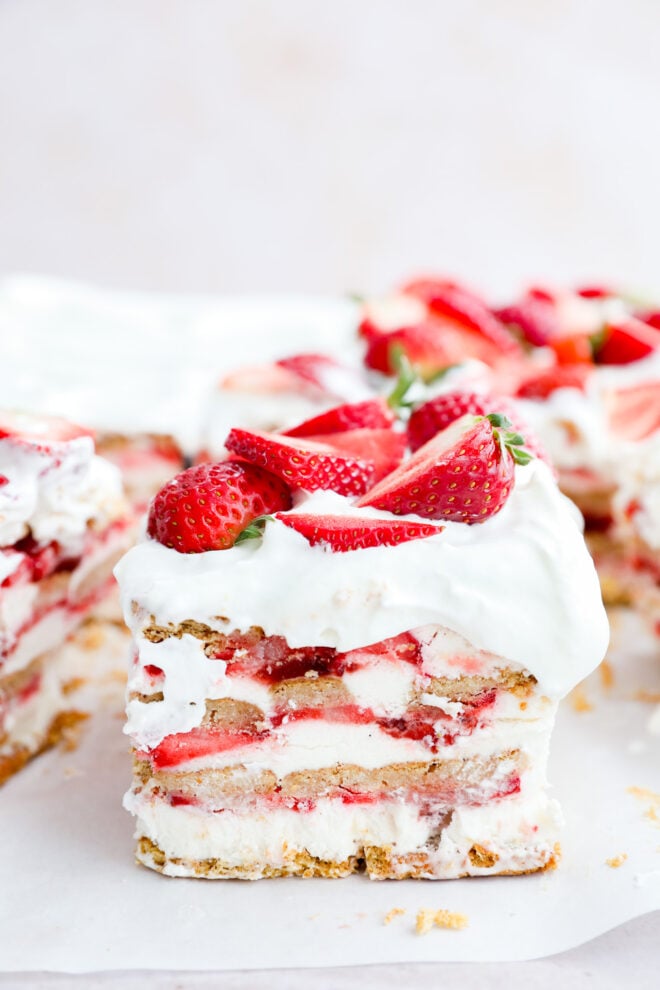 A slice of strawberry icebox cake with layers of whipped cream, graham crackers, and fresh strawberries. The slice is topped with halved strawberries, while more strawberry icebox cake is visible in the background.