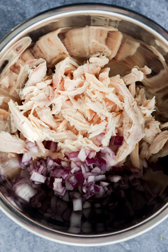 A metal bowl containing shredded cooked chicken and chopped red onions, ready to be mixed