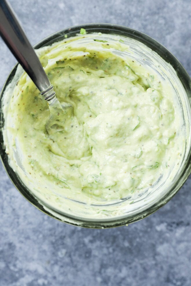 A glass bowl filled with creamy, light green avocado dressing, flecked with fresh herbs, sits on a gray textured surface with a spoon resting inside.