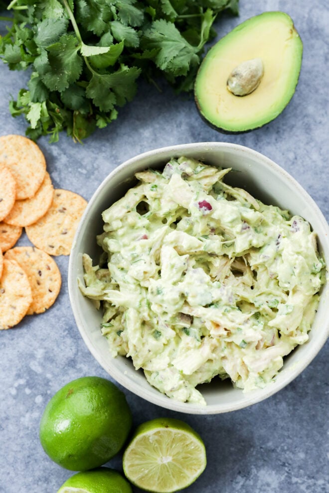 A bowl of creamy avocado chicken salad guacamole with pieces of chicken, surrounded by fresh cilantro, half an avocado, lime halves, and round crackers on a gray surface.