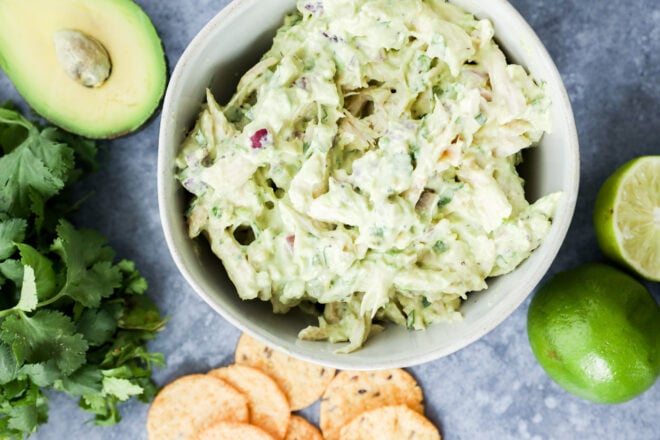 A bowl of creamy avocado chicken salad sits on a gray surface, surrounded by an avocado half, fresh cilantro, a halved lime, a whole lime, and round crackers&mdash;perfect for enjoying this flavorful avocado chicken salad.
