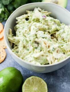 A bowl of creamy lunch sits on a gray surface, surrounded by a halved lime, whole lime, fresh cilantro, an avocado, and round crackers.
