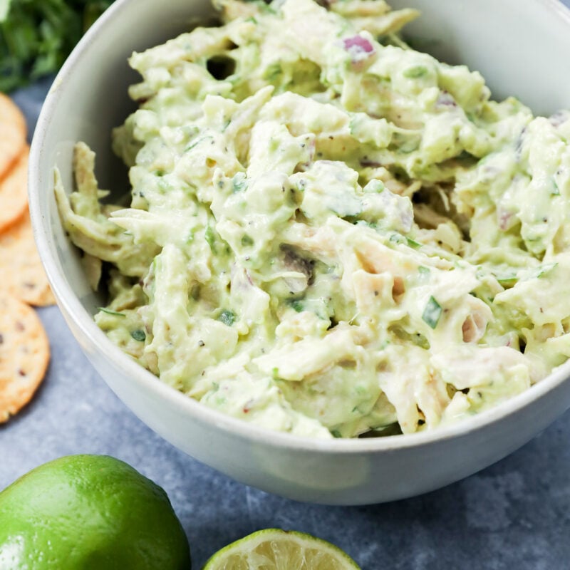 A bowl of creamy lunch sits on a gray surface, surrounded by a halved lime, whole lime, fresh cilantro, an avocado, and round crackers.