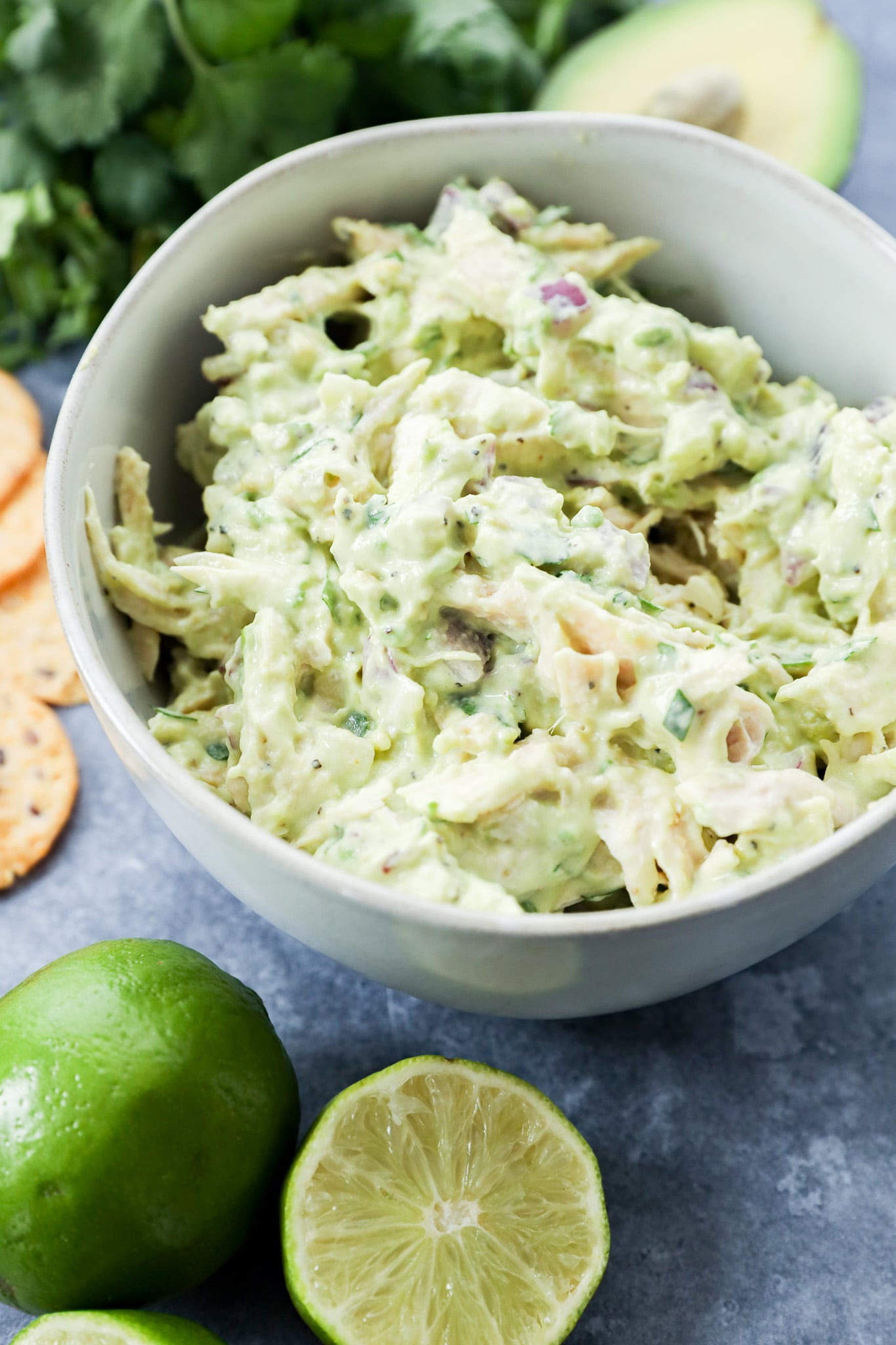 A bowl of creamy lunch sits on a gray surface, surrounded by a halved lime, whole lime, fresh cilantro, an avocado, and round crackers.
