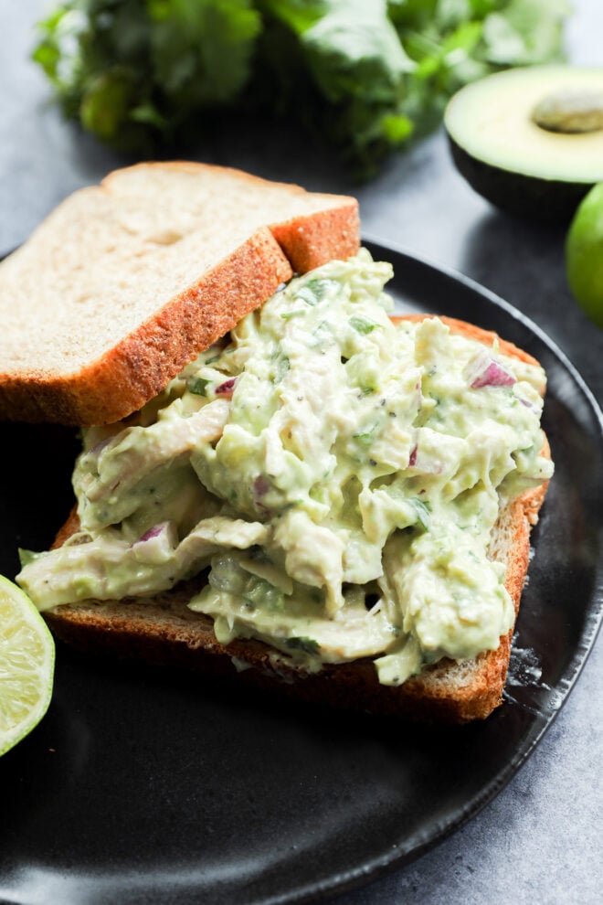 A close-up of an open-faced sandwich featuring avocado chicken salad on whole wheat bread, garnished with fresh cilantro, with lime and avocado halves in the background.