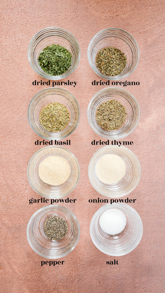 Eight small glass bowls on a pink surface, each containing a different garlic herb seasoning: dried parsley, oregano, basil, thyme, garlic powder, onion powder, pepper, and salt. Labels clearly identify each seasoning.