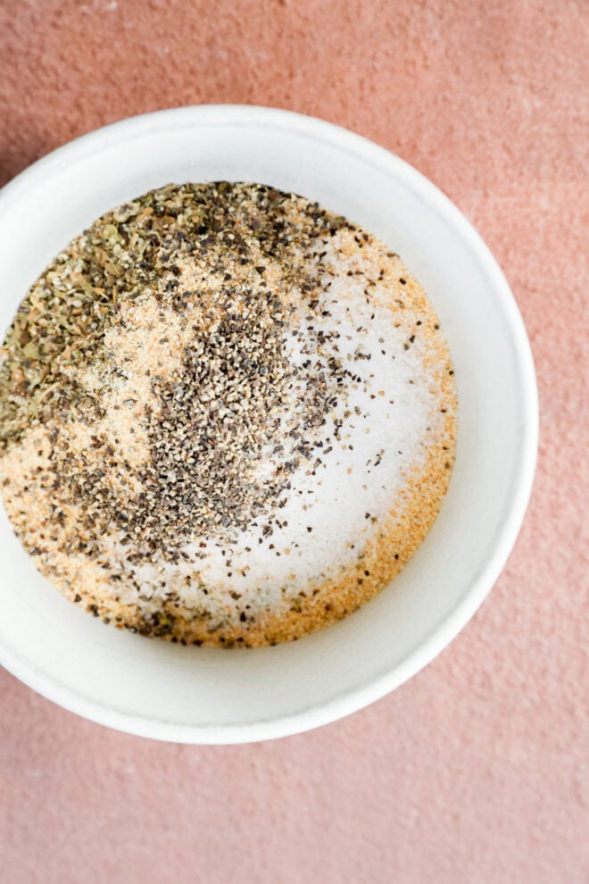 A white bowl on a pink surface contains spices, featuring a blend of salt, dried herbs, ground black pepper, and garlic powder for flavorful seasoning.