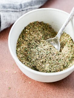 A white bowl filled with mixed garlic herb seasoning, with a small silver spoon resting inside. A textured cloth is partially visible in the background on a brown surface.
