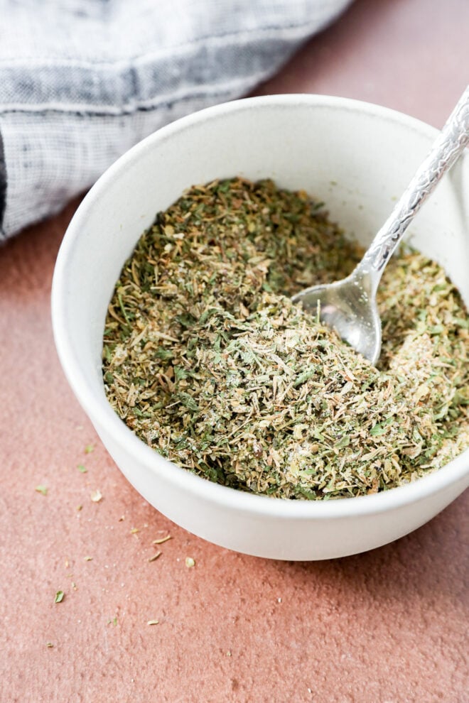 A white bowl filled with mixed garlic herb seasoning, with a small silver spoon resting inside. A textured cloth is partially visible in the background on a brown surface.