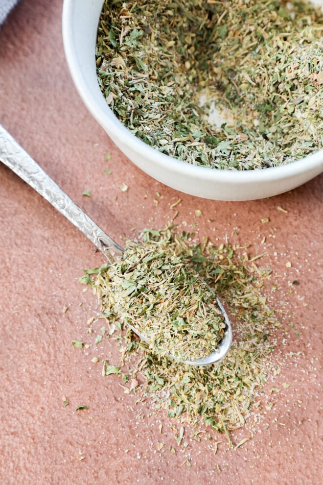 A close-up of a spoonful of dried mixed herbs, spilling onto a brown surface, with a small white bowl filled with more of the herb mix in the background.