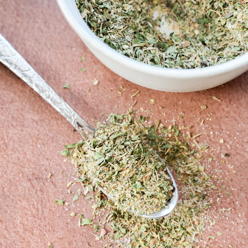 A close-up of a spoonful of dried mixed herbs, spilling onto a brown surface, with a small white bowl filled with more of the herb mix in the background.