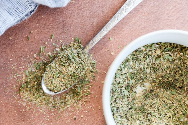 A close-up of a spoonful of garlic herb seasoning, with some herbs scattered on a pink surface next to a white bowl filled with more dried herbs. A piece of light-colored cloth is visible in the corner.