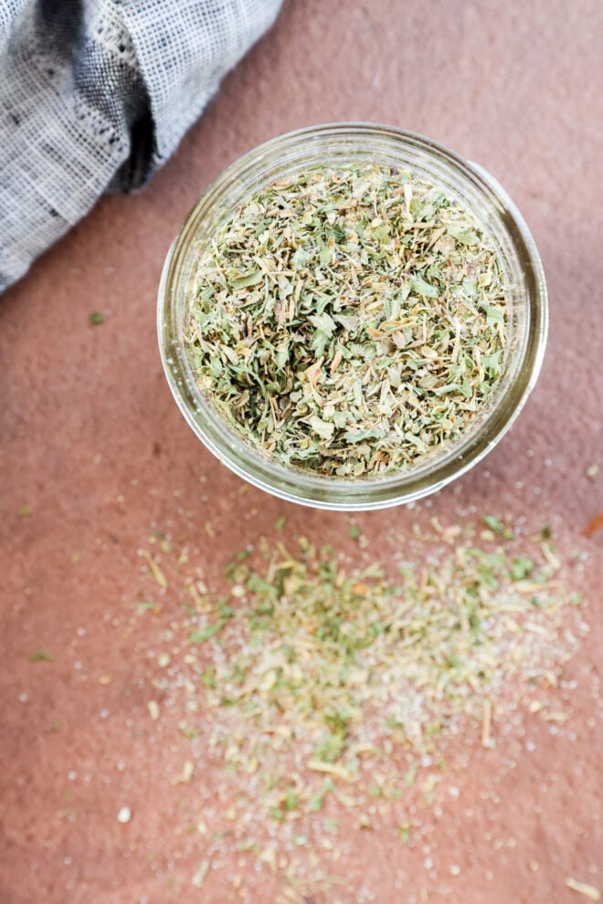 A jar filled with dried garlic herb seasoning sits on a brown surface, with some of the savory mixture spilled beside it. A textured grey cloth is partially visible in the upper left corner.