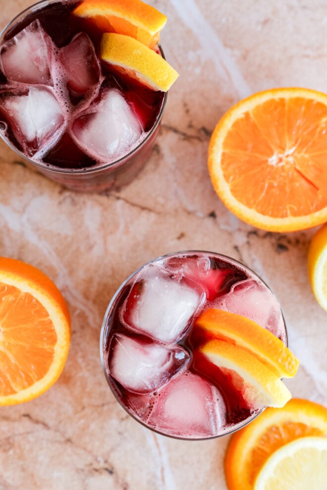 Two glasses filled with a red wine cocktail, garnished with orange slices, sit on a marble surface. Halved oranges are placed around the glasses.