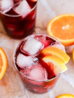 A glass filled with a dark red wine cocktail, ice cubes, and orange slices as garnish sits surrounded by fresh orange slices on a light surface. Another similar glass is partially visible in the background.