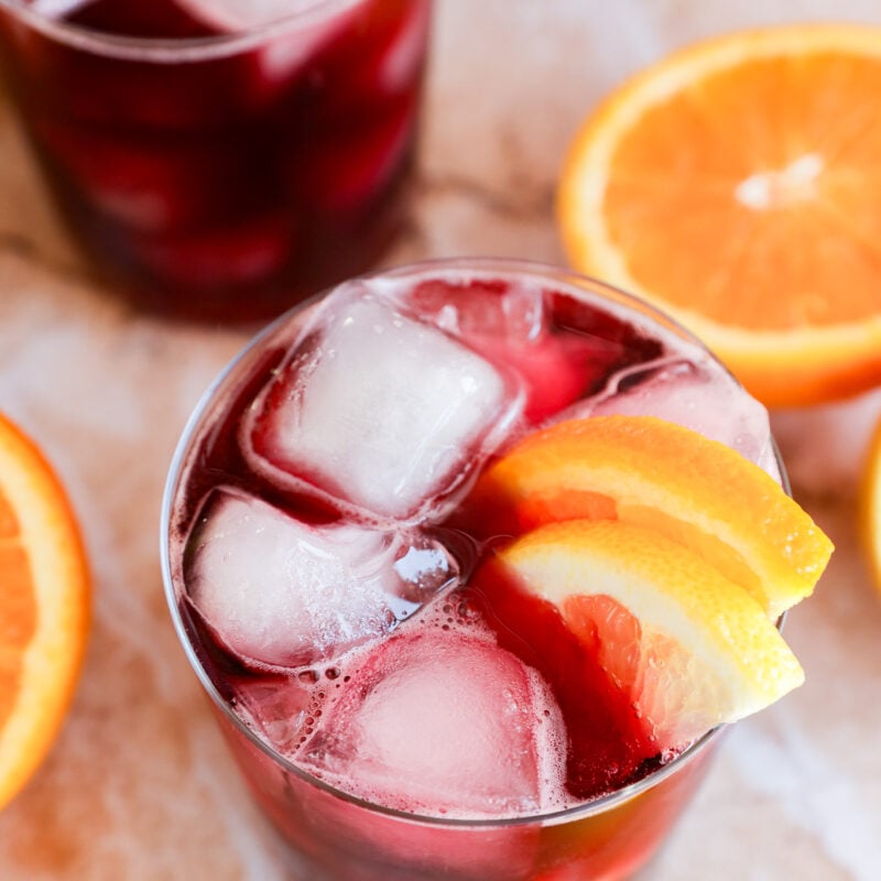 A glass filled with a dark red wine cocktail, ice cubes, and orange slices as garnish sits surrounded by fresh orange slices on a light surface. Another similar glass is partially visible in the background.