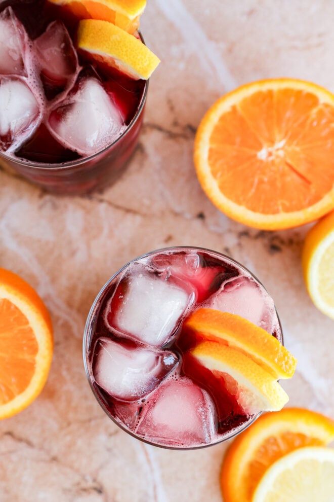 Two glasses of a dark purple red wine cocktail with ice cubes and orange slices, placed on a light marble surface, surrounded by fresh orange slices.