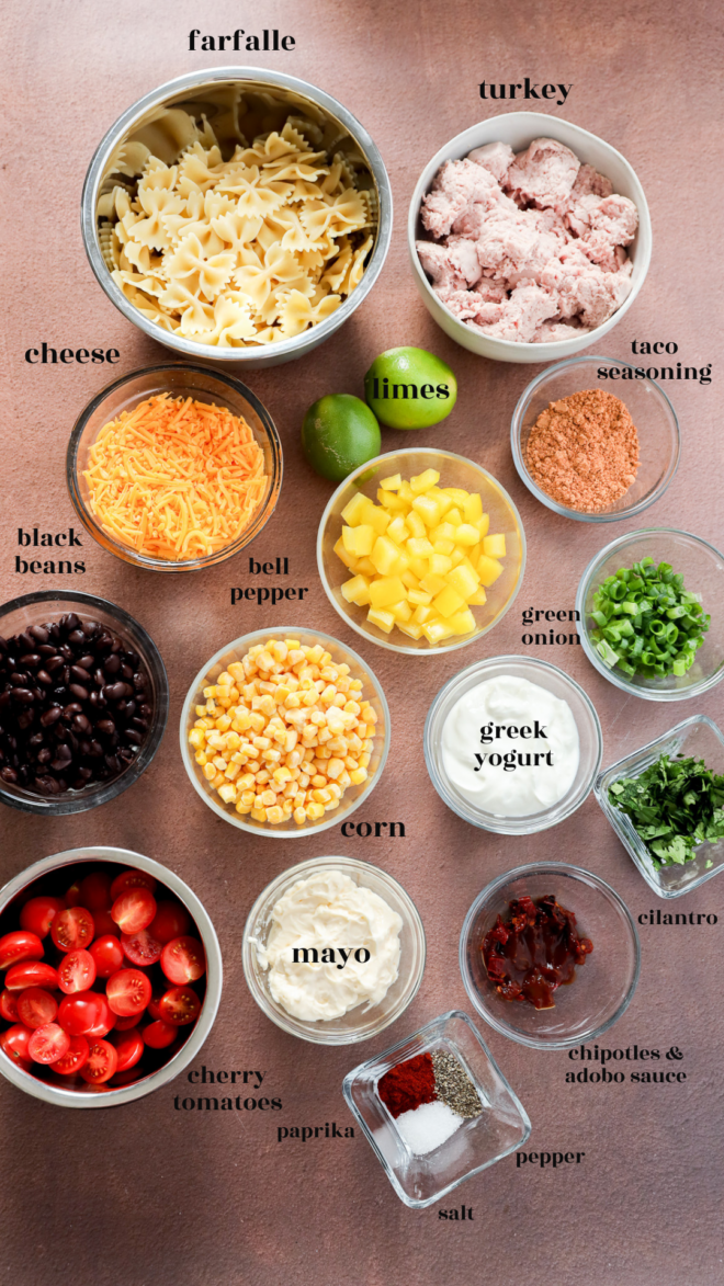 A top-down view of bowls containing labeled ingredients for Taco Pasta Salad: farfalle pasta, turkey, taco seasoning, cheese, black beans, bell pepper, green onion, limes, greek yogurt, mayo, corn, cilantro, cherry tomatoes, chipotles & adobo sauce, paprika, salt, and pepper.