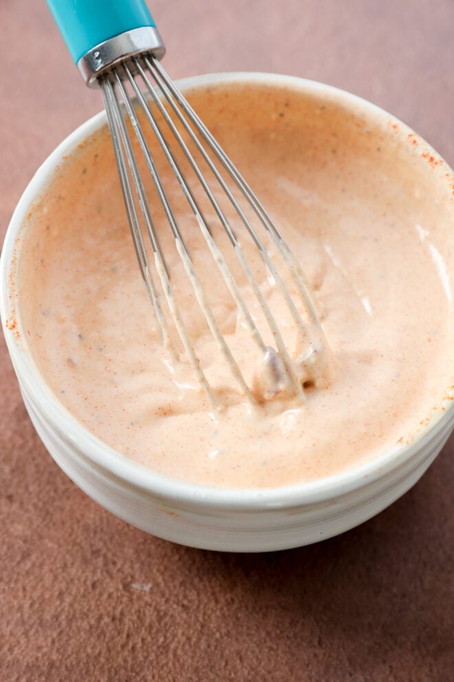 A metal whisk with a turquoise handle mixing a creamy, orange-colored sauce in a white bowl on a brown surface.