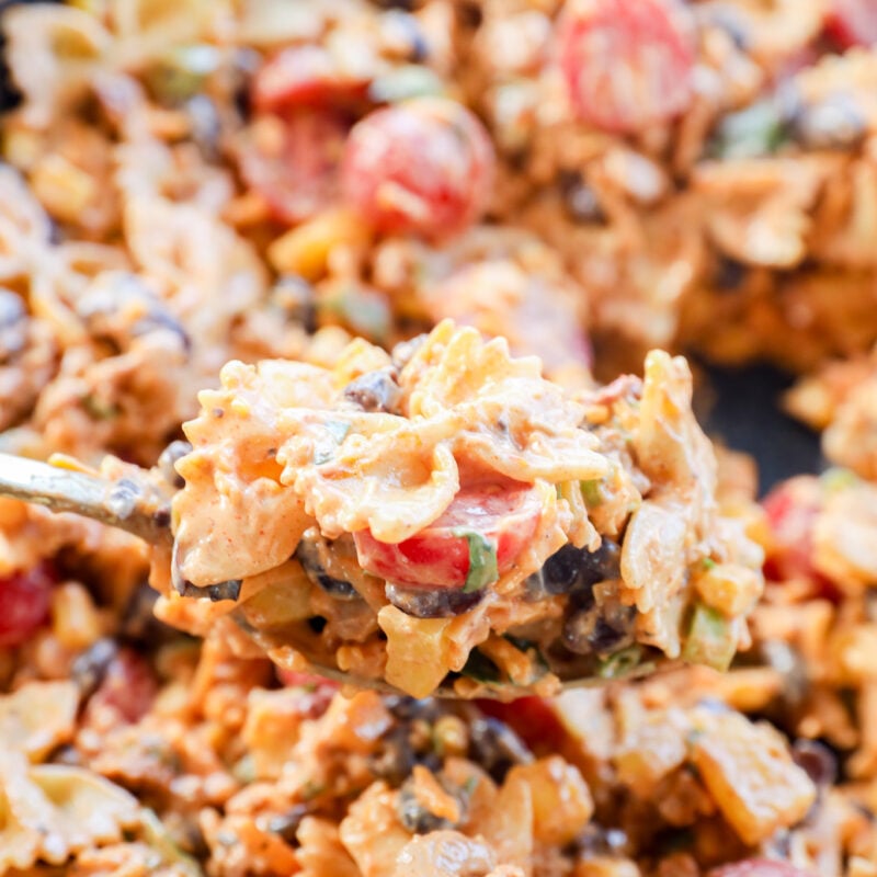 A close-up of a spoonful of Taco Pasta Salad with creamy bowtie noodles, cherry tomatoes, black beans, green onions, and ground meat, with more of the flavorful dish blurred in the background.