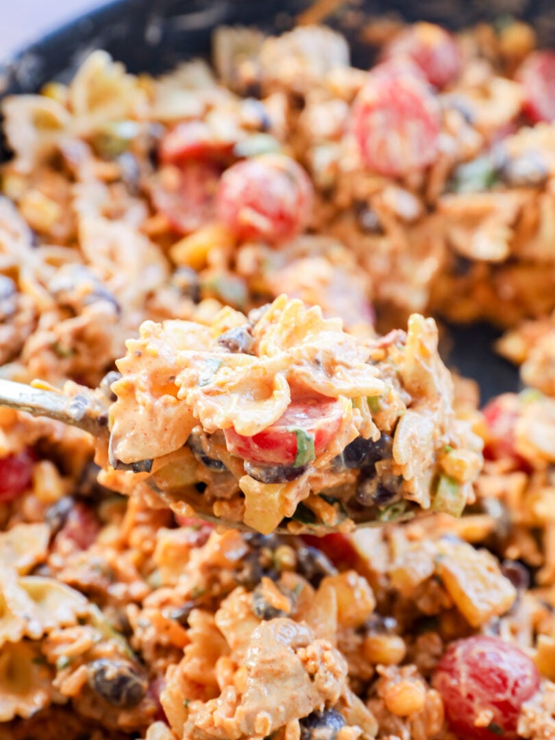 A close-up of a spoonful of Taco Pasta Salad with creamy bowtie noodles, cherry tomatoes, black beans, green onions, and ground meat, with more of the flavorful dish blurred in the background.