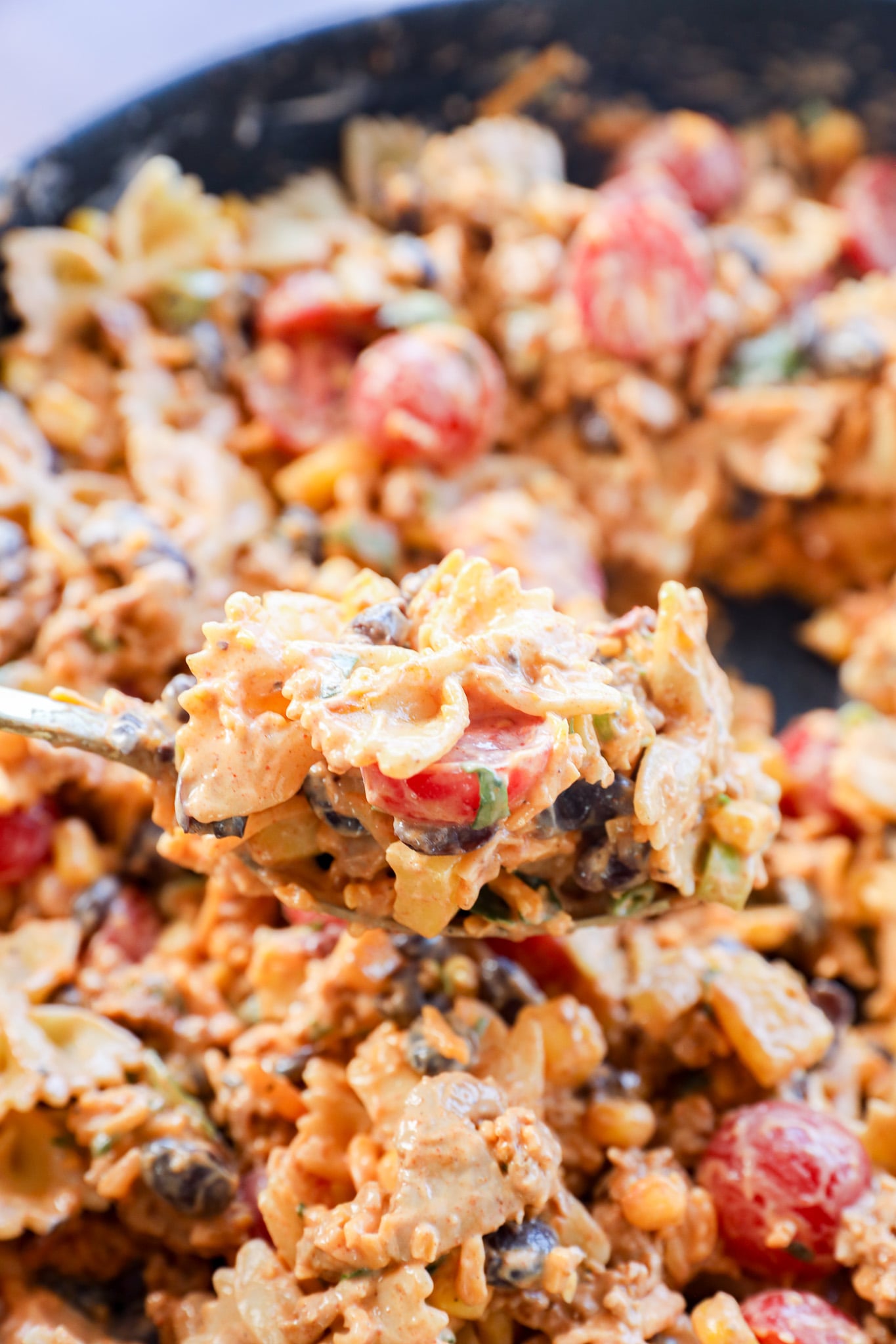 A close-up of a spoonful of Taco Pasta Salad with creamy bowtie noodles, cherry tomatoes, black beans, green onions, and ground meat, with more of the flavorful dish blurred in the background.