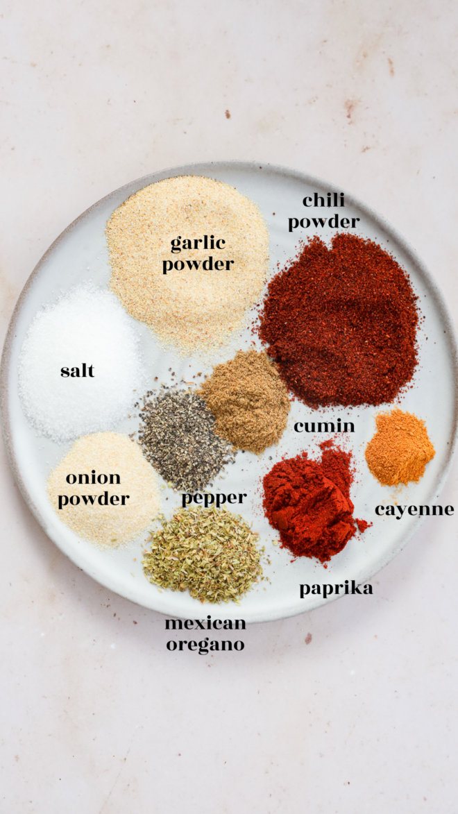A white plate with labeled piles of seasonings, including chili powder, garlic powder, cumin, cayenne, paprika, Mexican oregano, pepper, onion powder, and salt—each essential for creating the perfect Carne Asada Seasoning—arranged neatly on a light surface.